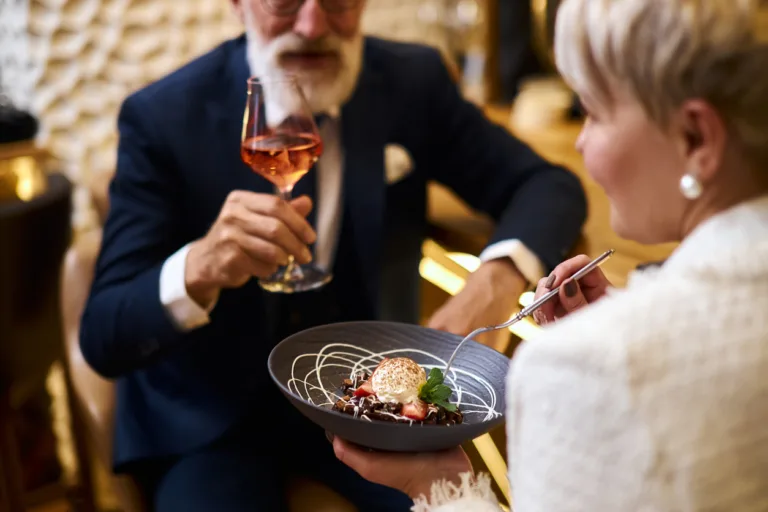 Close image of hand with glass of champagne, sweet dessert on grey dish. Beared man in tuxedo drinking