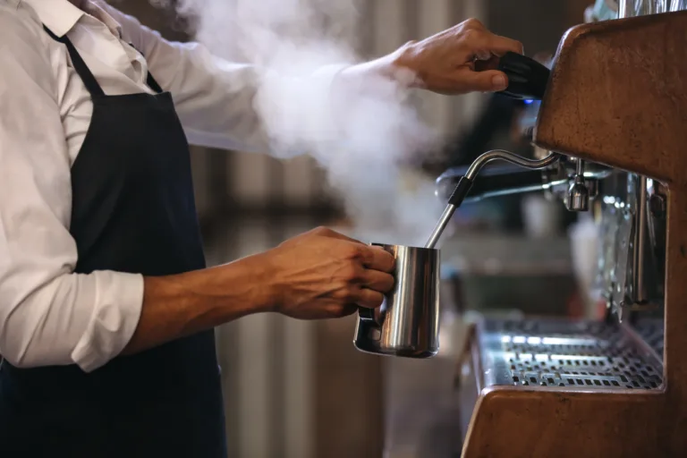 Coffee shop worker preparing coffee on steam espresso coffee machine. Cropped shot of man working in coffee shop wearing an apron.