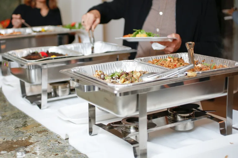 A view of several chafer dishes filled with savory entrees, seen at a local catered event. Guests are seen serving themselves.