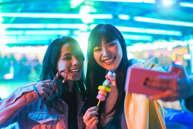 Happy asian girls eating candy sweets and taking selfie at amusement park - Young trendy friends having fun with technology trend - Tech, friendship and influencer concept - Focus on right female face