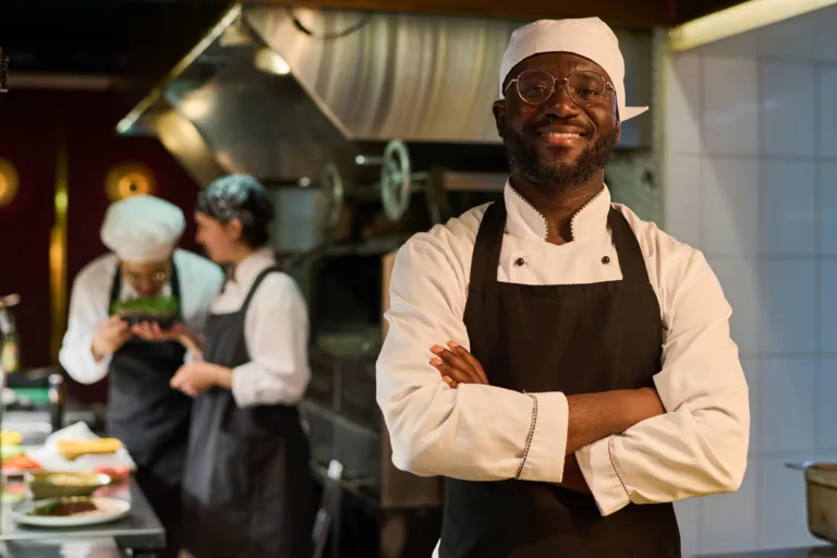 Happy African American male chef in uniform standing in front of camera in restaurant kitchen and keeping his arms crossed by chest restaurant server uniforms
